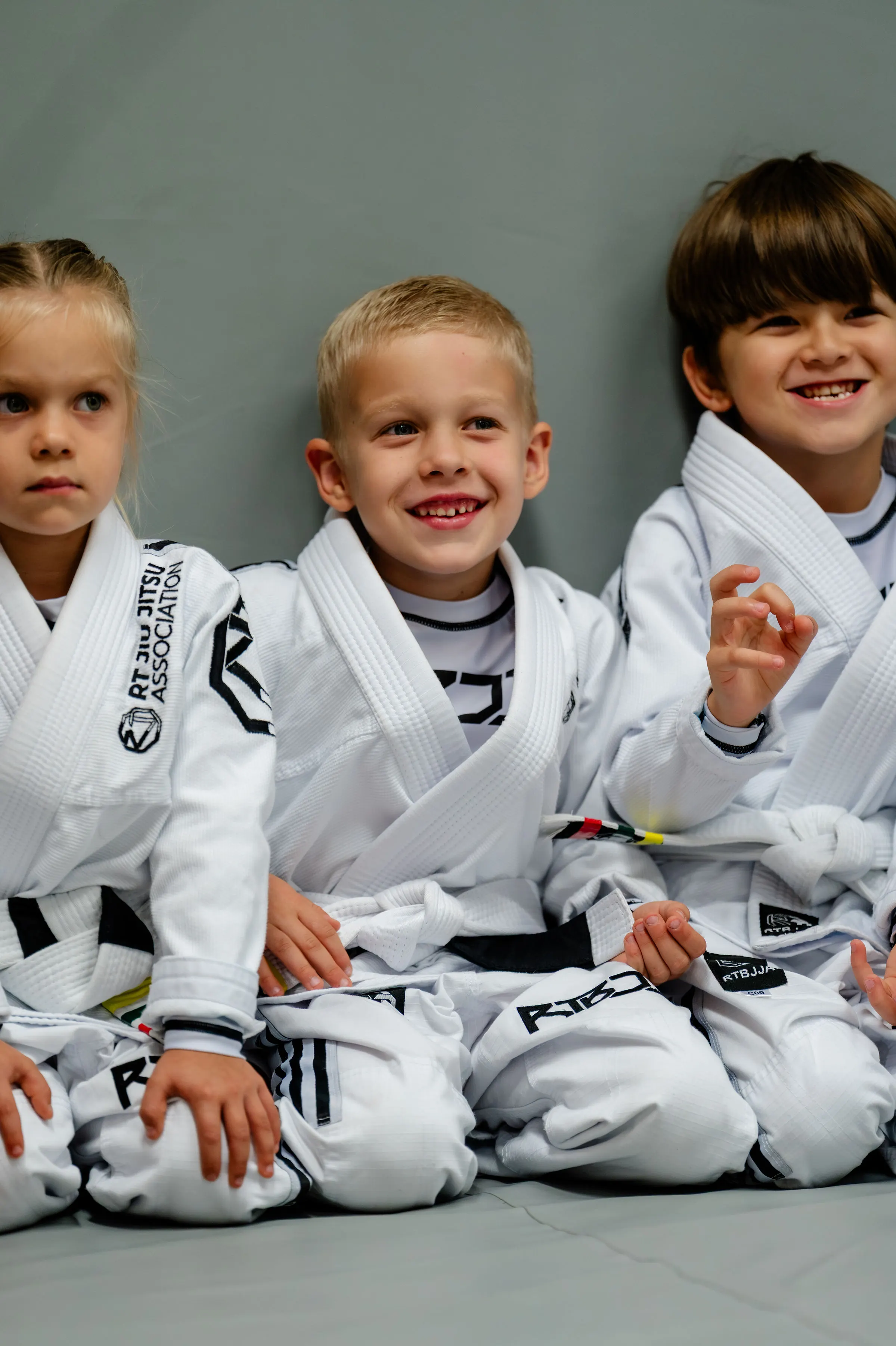 Kids Jiu Jitsu class at RT Beachside: three children in white Gis sitting against the wall, laughing