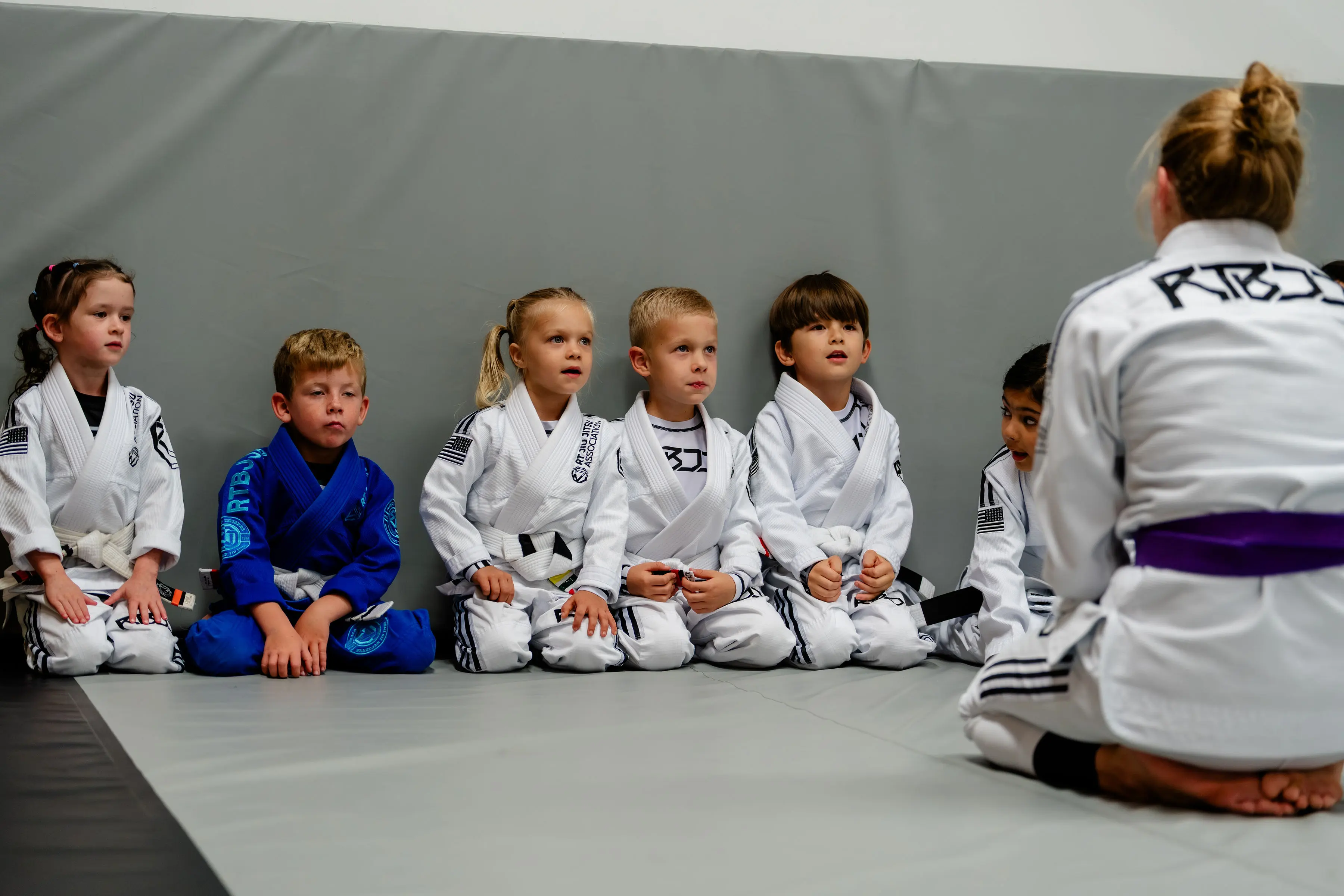 Kids Jiu Jitsu class at RT Beachside: six children in Gis sitting against the wall, listening to the instructor in front of them