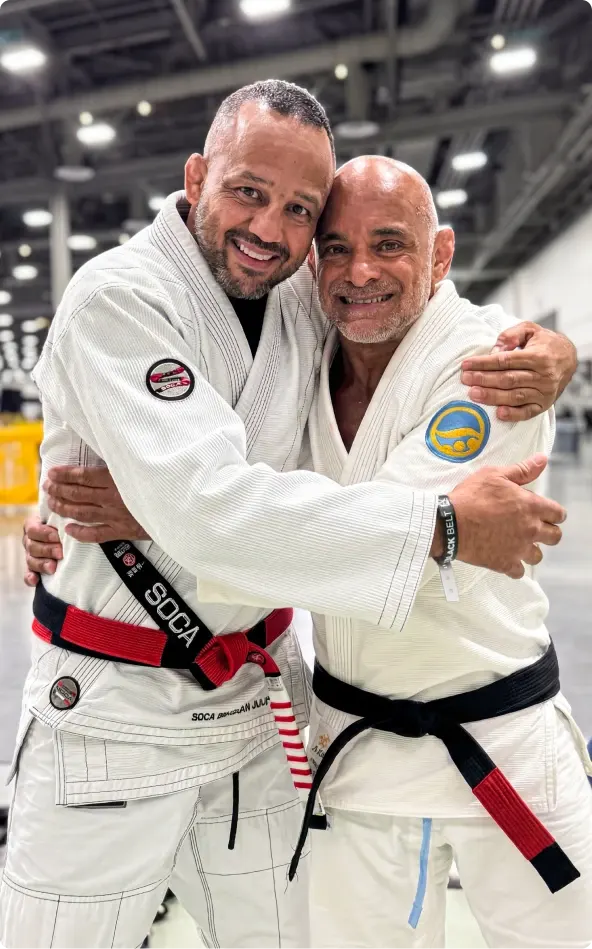 Master Renato Tavares in a white Gi, smiling and hugging a fellow black belt on the mats